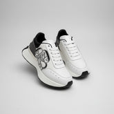 White sneakers with black accents held by a gloved hand, with Alexander McQueen boxes in the background.